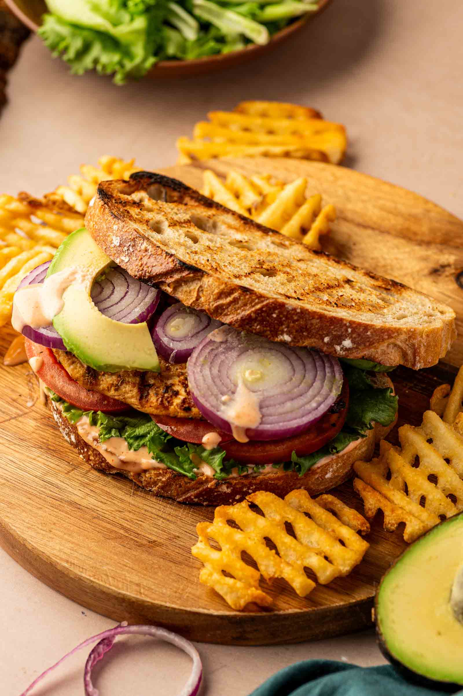 A grilled chicken sandwich with lettuce, tomato, red onion, and avocado on toasted bread, served with waffle fries on a wooden board.