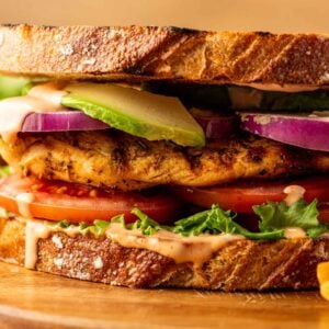 A close-up of a grilled chicken sandwich with lettuce, tomato, red onion, avocado, and sauce on toasted bread, served on a wooden board.