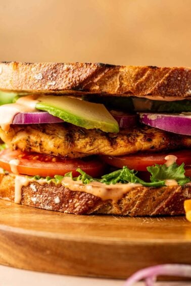 A close-up of a grilled chicken sandwich with lettuce, tomato, red onion, avocado, and sauce on toasted bread, served on a wooden board.