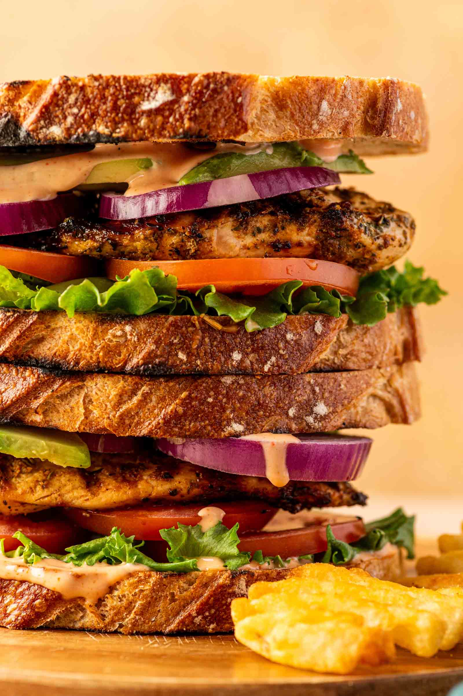Close-up of a stacked grilled chicken sandwich with lettuce, tomato, red onion, sauce, and toasted bread, served with a side of potato chips.