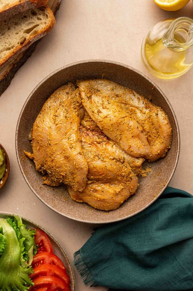 A bowl of seasoned raw chicken breasts sits on a counter beside sliced bread, a bottle of oil, lemon, and plates of sliced lettuce, tomato, and pickles.