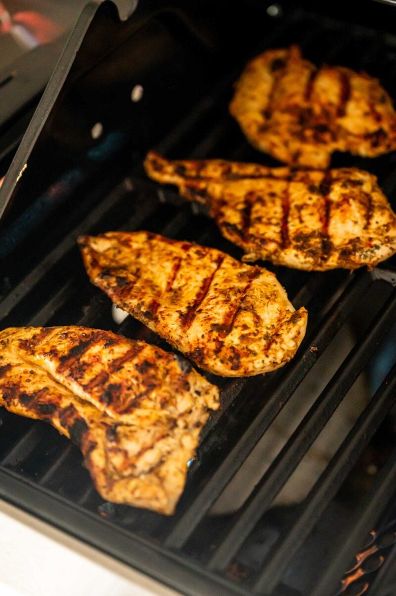 Four seasoned chicken breasts cooking on a grill, with visible grill marks on the meat.