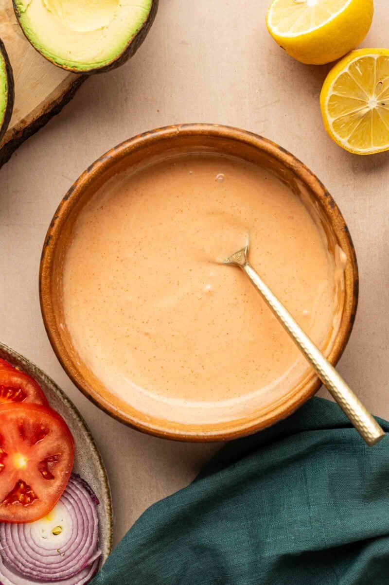 A bowl of creamy pink Spicy Mayo sauce with a spoon inside, surrounded by sliced tomato, red onion, avocado, and lemon halves on a beige surface.