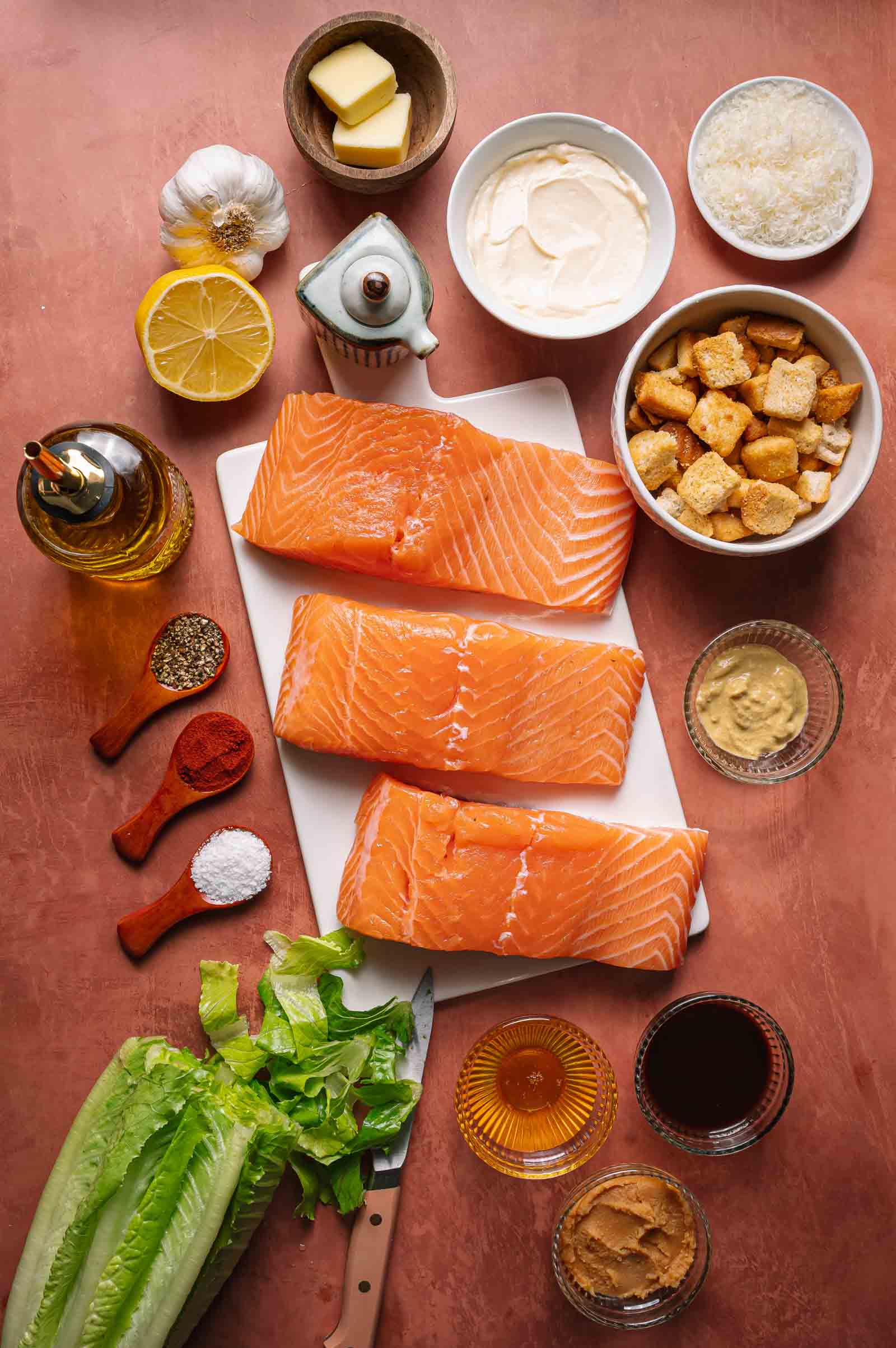 Salmon fillets on a cutting board surrounded by lettuce, croutons, condiments, spices, garlic, lemon, butter, grated cheese, and salad dressing ingredients on a countertop.