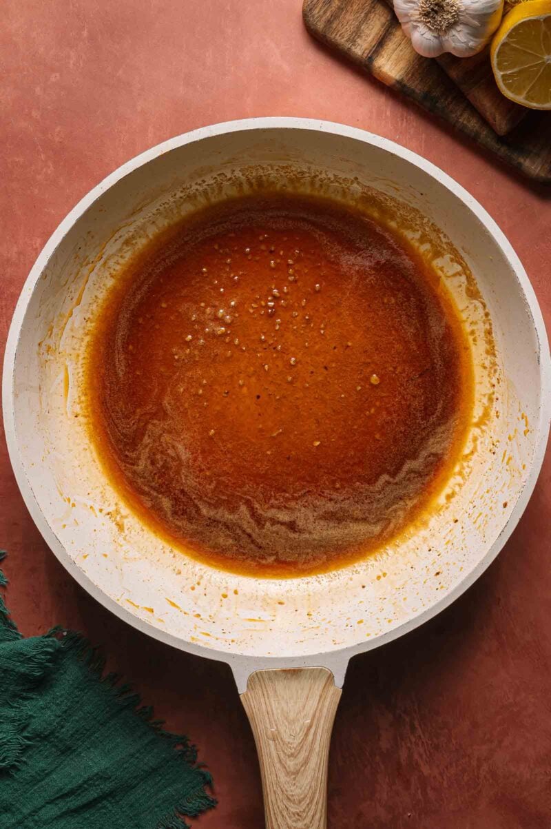A white skillet with a hot honey sauce simmering inside, placed on a brown surface next to a green cloth and a wooden board with garlic and lemon slices.