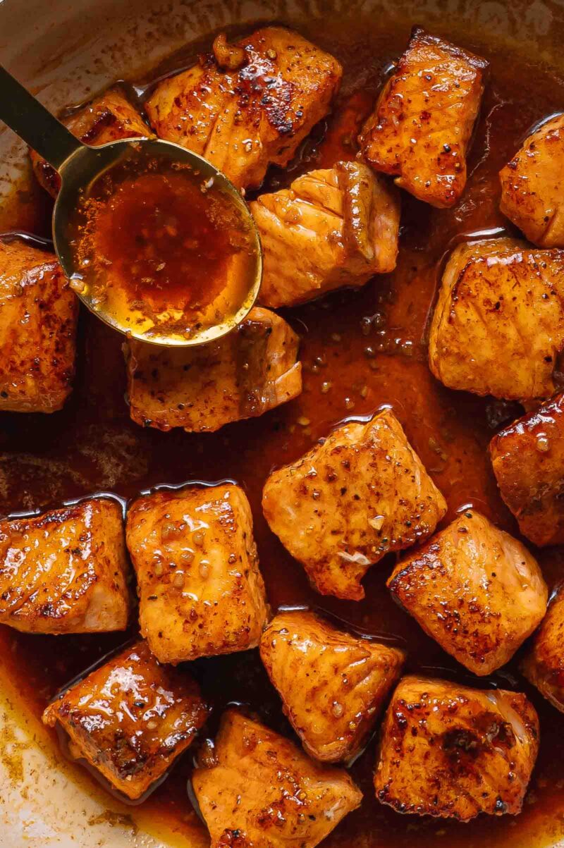 Close-up of cooked salmon bites coated in a glossy hot honey sauce, with a spoon drizzling more sauce over the pieces.