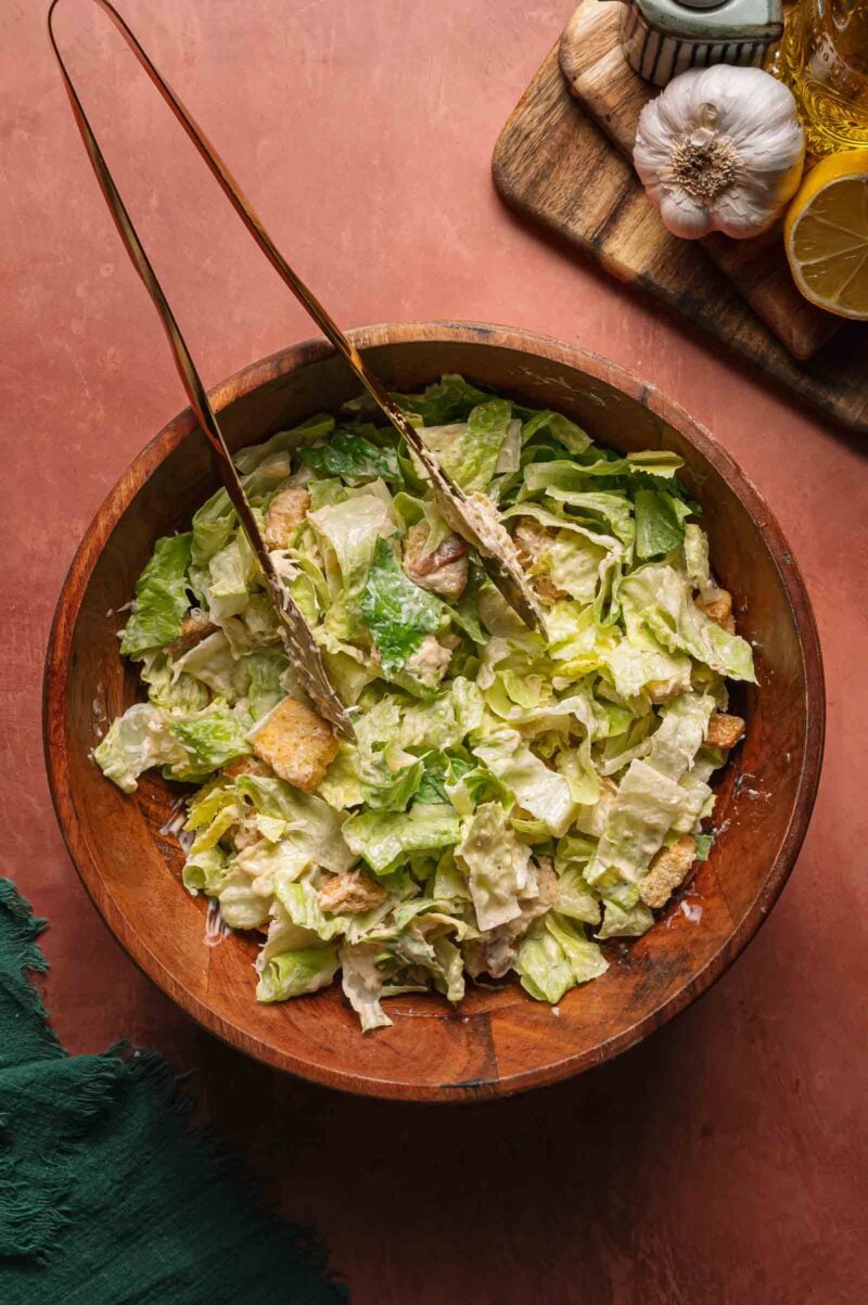 A wooden bowl filled with Caesar salad, filled with romaine lettuce, croutons, and creamy dressing, with metal tongs resting on top. Garlic, lemon, and olive oil are visible nearby.