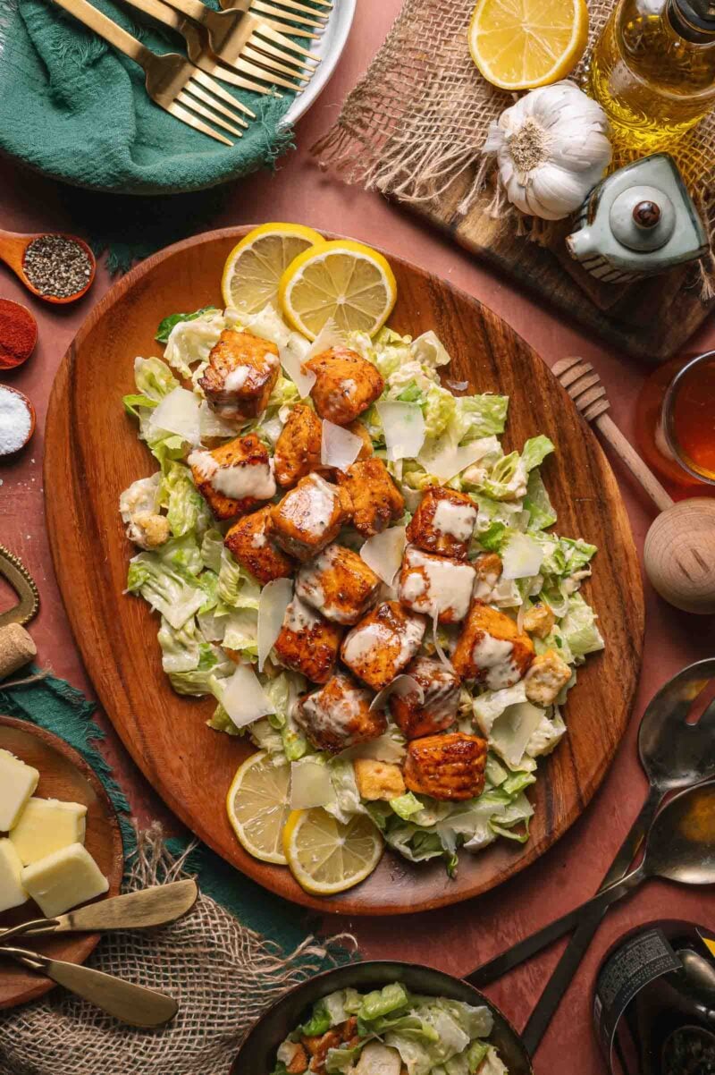 A wooden platter with hot honey salmon Caesar salad, topped with shaved parmesan and Caesar dressing, garnished with lemon slices. Various utensils and seasonings surround the platter.