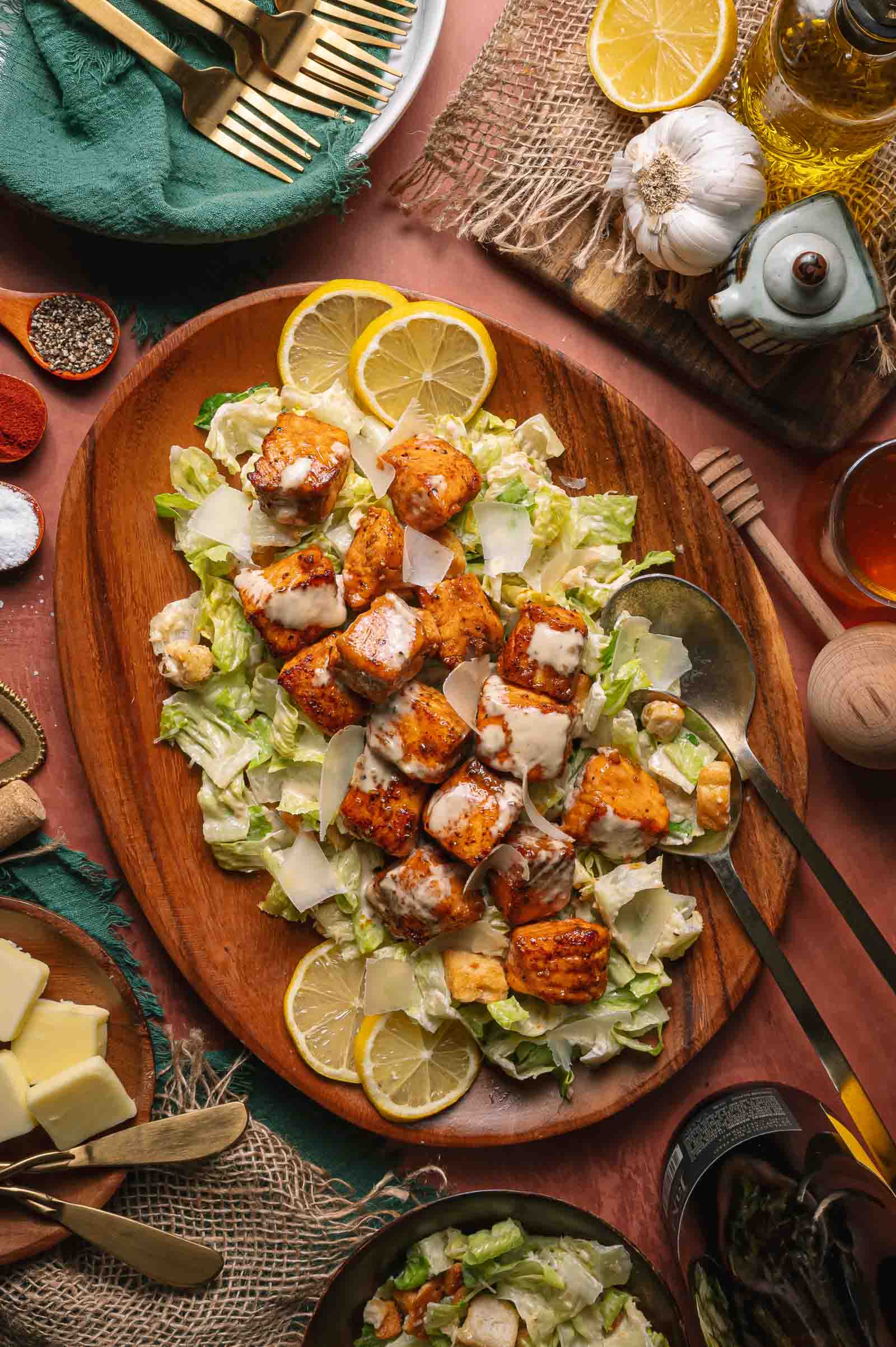 Hot Honey Salmon Caesar salad topped with shaved Parmesan and lemon slices on a wooden platter, surrounded by various ingredients and utensils.
