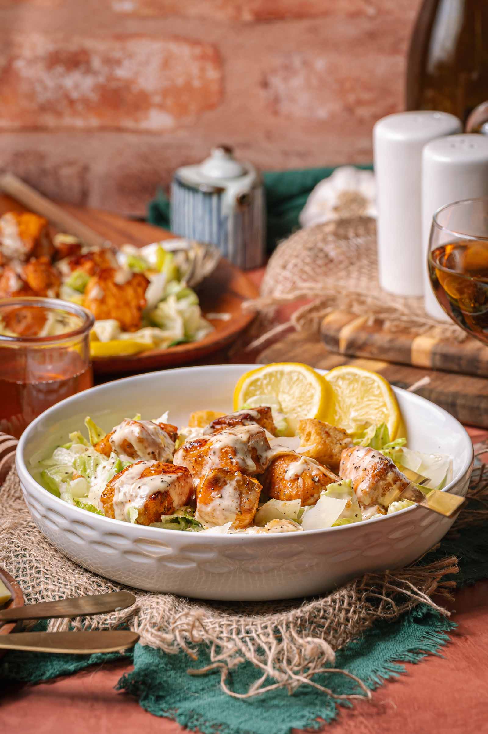 A white bowl filled with hot honey salmon caesar salad. A drink and other dishes are visible in the background.