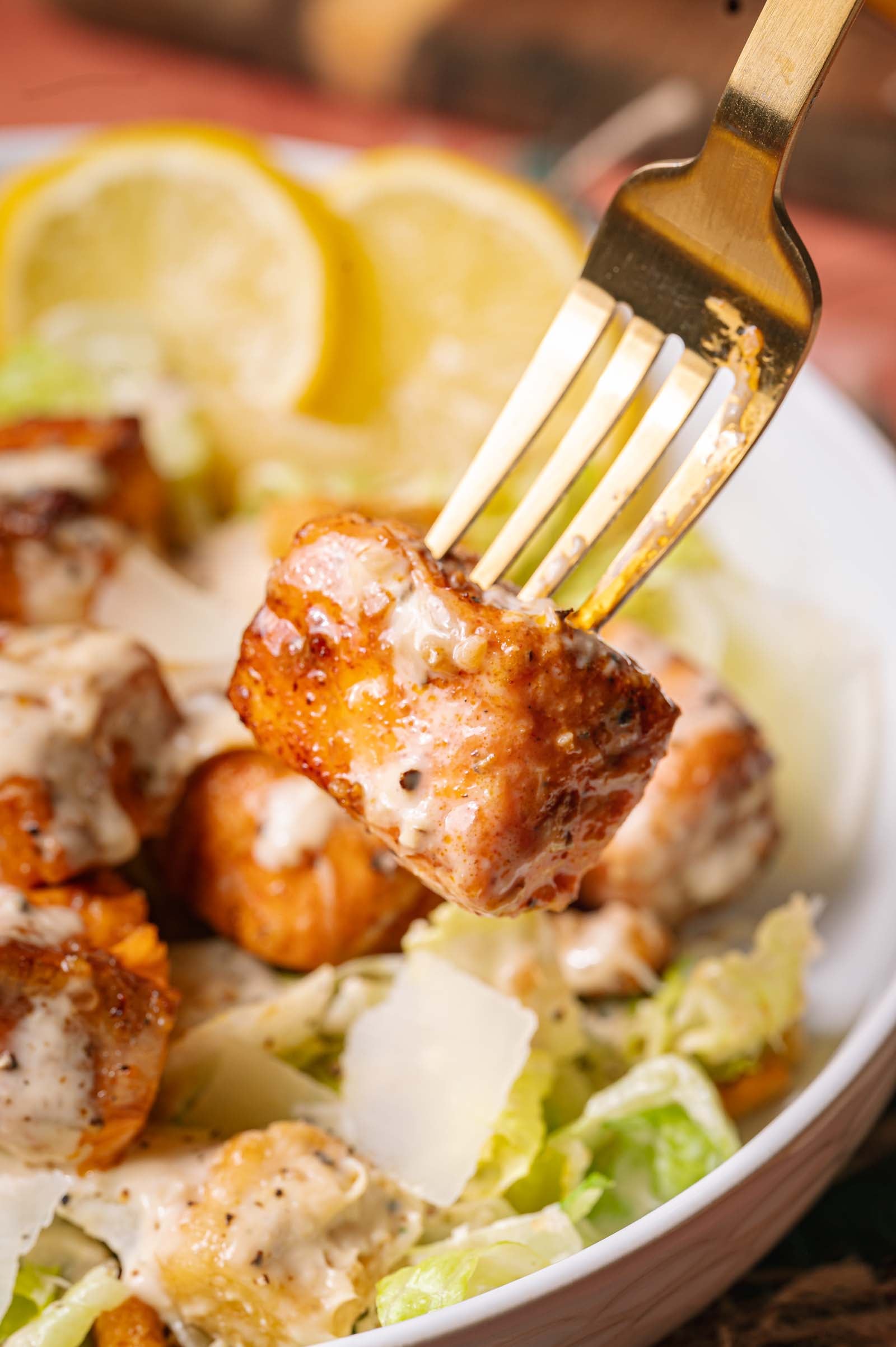A close-up of a fork holding a hot honey salmon bite above a Caesar salad with lettuce, Parmesan shavings, dressing, and lemon slices in the background.
