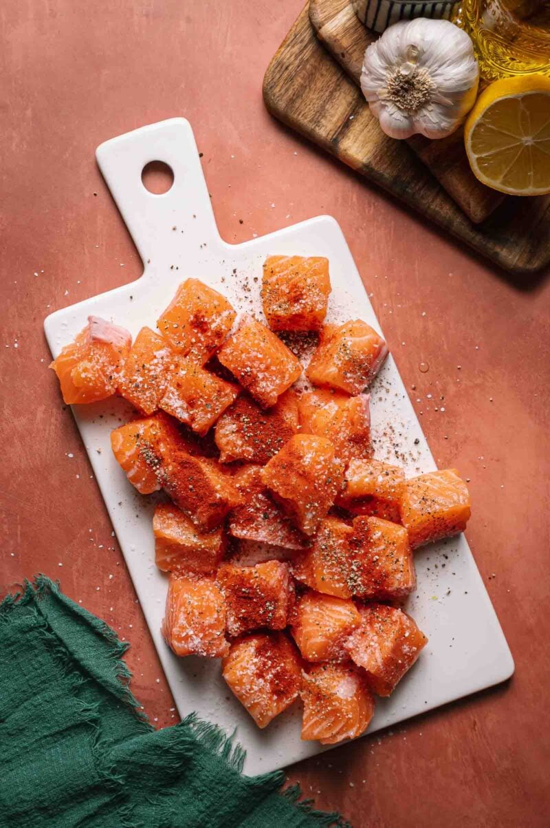 Raw salmon cubes seasoned with salt and pepper on a white cutting board, with garlic, lemon, and oil visible on a wooden board nearby.