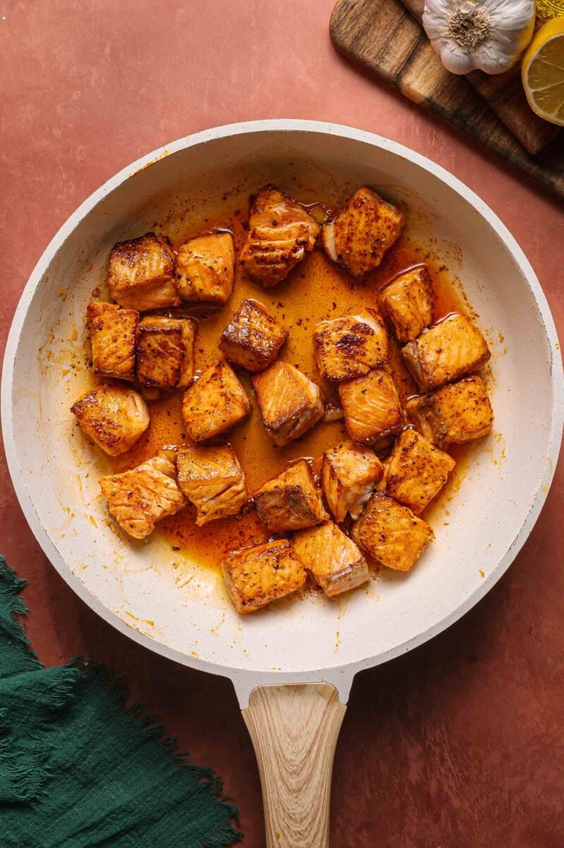 Cubes of cooked salmon in a white skillet with a seasoned sauce, next to a green cloth, a lemon slice, and garlic.