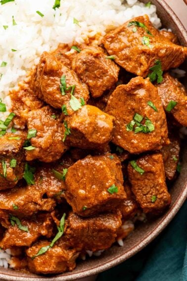 A close-up of lamb vindaloo with red sauce and chopped cilantro served over white rice in a bowl.