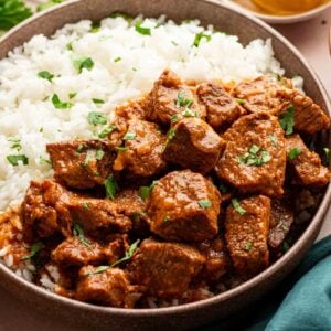A bowl of white rice topped with chunks of saucy, lamb vindaloo, garnished with chopped cilantro, with lemon wedges and fresh cilantro on the side.