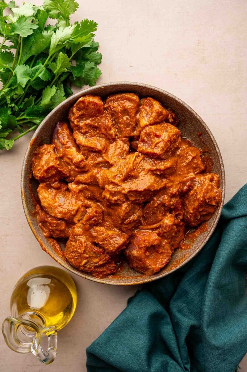 A bowl of marinated lamb pieces in reddish-brown sauce, with a bunch of fresh cilantro, a bottle of oil, and a teal cloth nearby on a light surface.