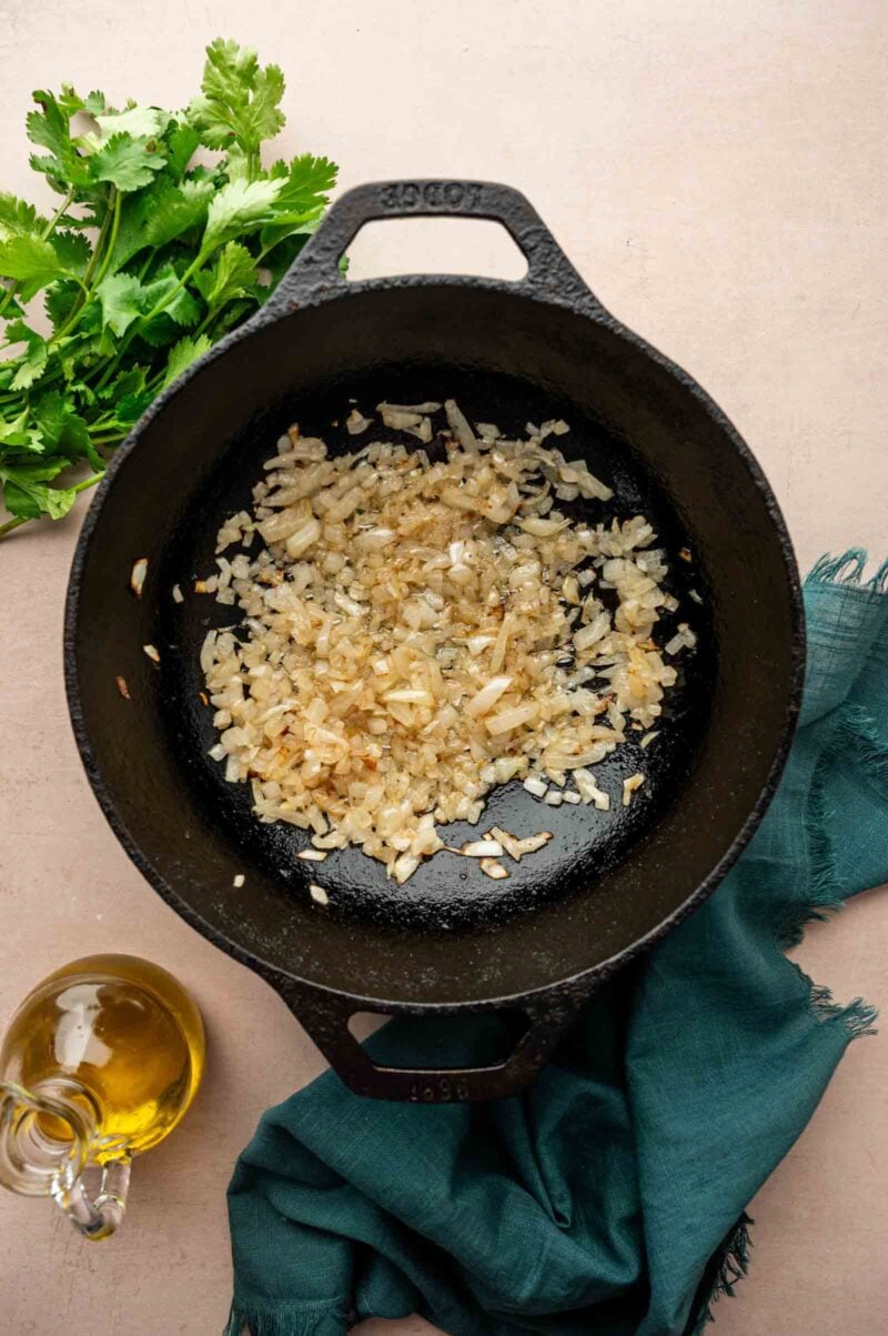 Chopped onions sautéing in a black cast iron pan, with a bunch of cilantro, a bottle of olive oil, and a green cloth nearby.