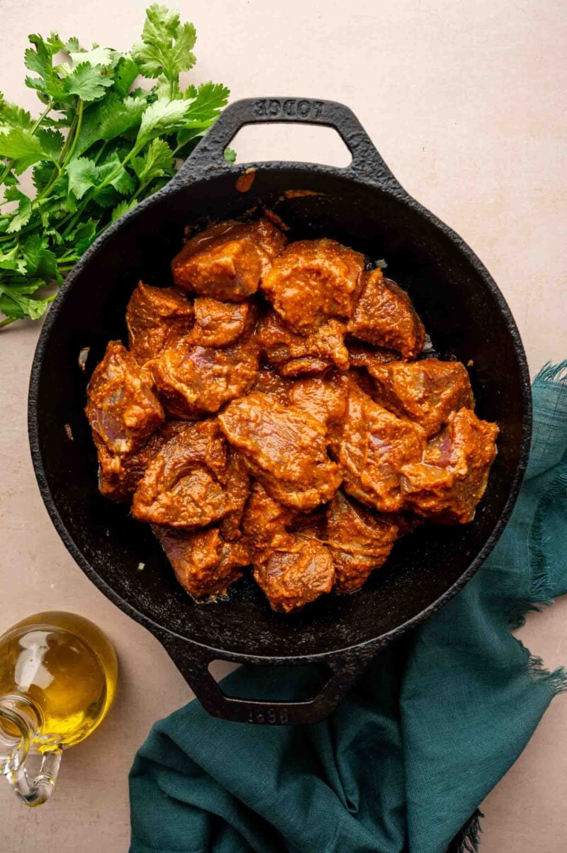 Chunks of marinated lamb in a black cast iron pot, with fresh cilantro, a bottle of oil, and a dark green cloth nearby on a light surface.