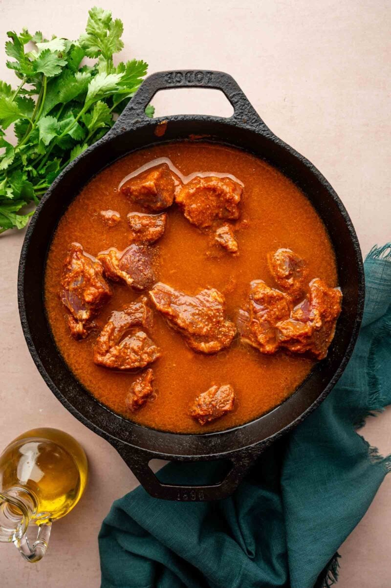 A black cast iron pot filled with chunks of lamb in a vindaloo sauce, placed on a beige surface with cilantro, a green cloth, and a bottle of oil nearby.