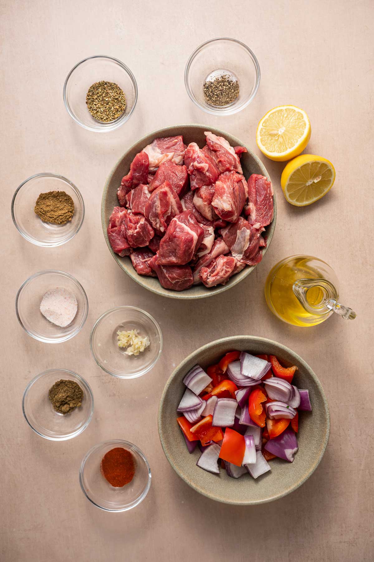 Bowl of raw lamb, bowl of chopped red onion and bell pepper, small bowls of spices, garlic, oil, and a halved lemon arranged on a light surface.