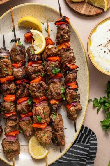 Grilled lamb and vegetable skewers with red peppers and onions on a plate, garnished with lemon wedges and fresh herbs, next to a bowl of white tzatziki sauce.