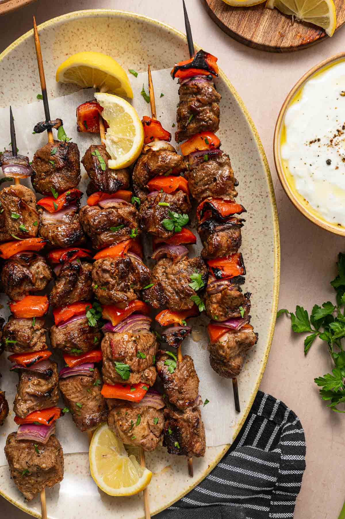 Grilled lamb and vegetable skewers with red peppers and onions on a plate, garnished with lemon wedges and fresh herbs, next to a bowl of white tzatziki sauce.