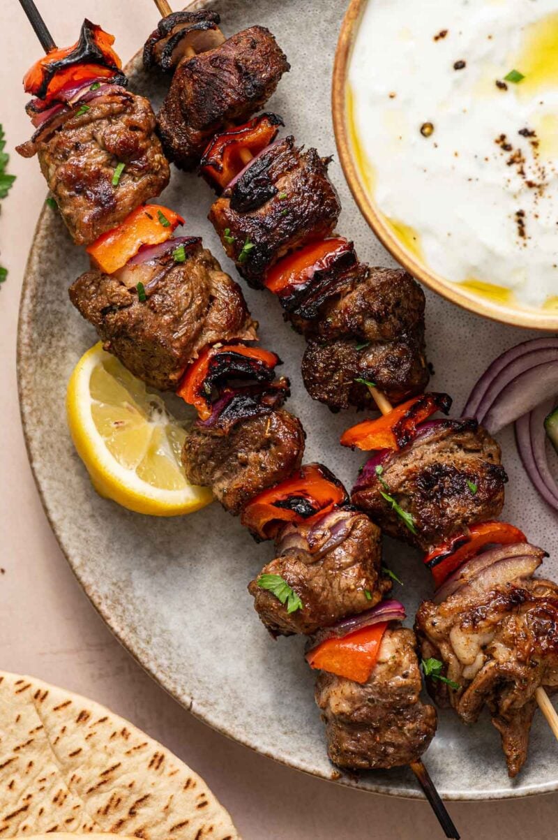 Two skewers of grilled lamb and vegetables on a plate with a lemon wedge, pita bread, sliced red onion, and a bowl of tzatziki sauce.
