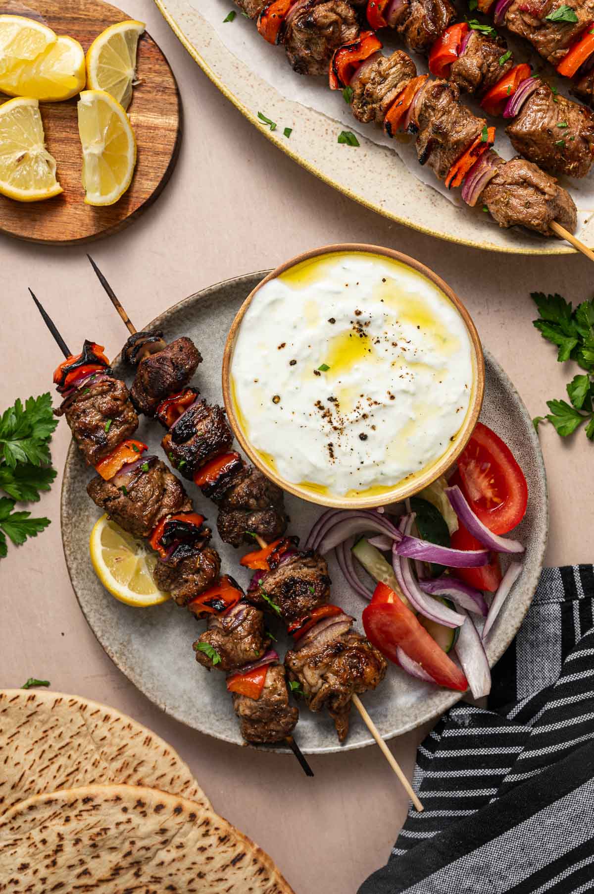 Two lamb kebab skewers with red bell peppers and onions served on a plate with dipping sauce, lemon wedges, tomato slices, red onion, pita bread, and fresh parsley.