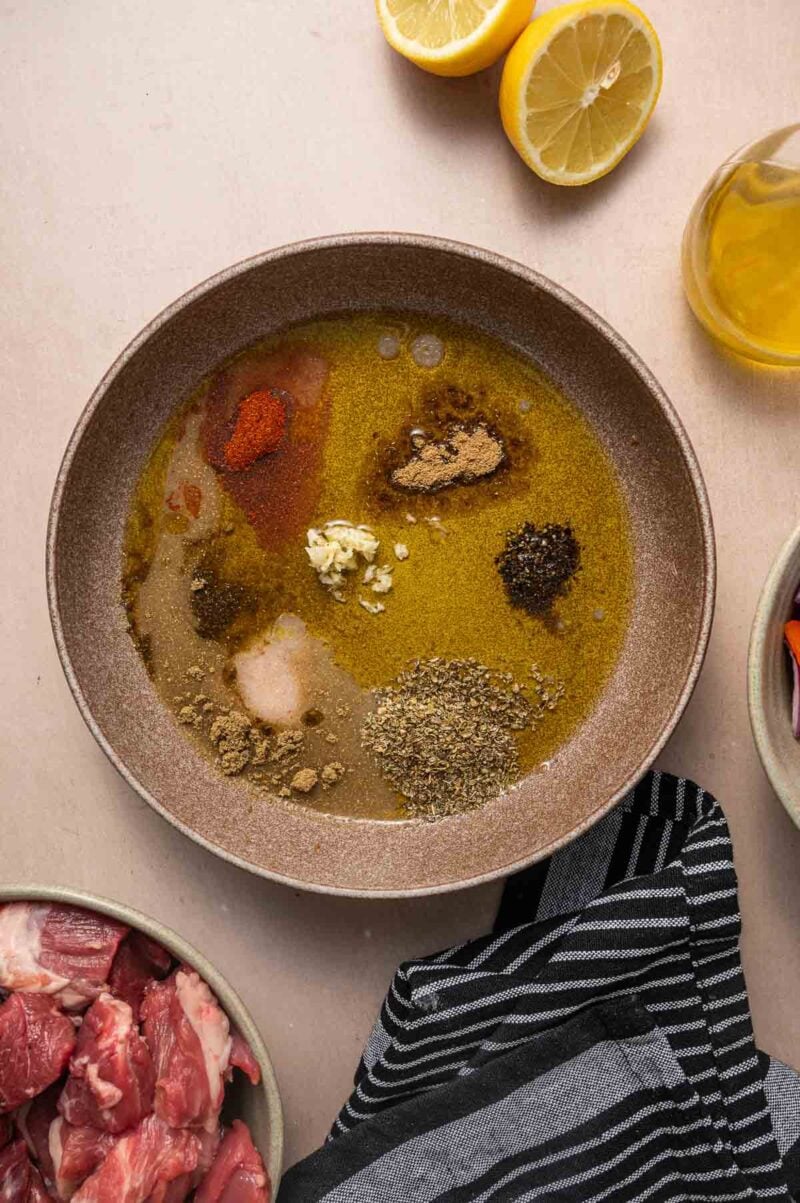 A bowl with various spices, minced garlic, olive oil, and lemon juice, surrounded by a bowl of raw meat, a striped towel, lemon halves, and a bottle of oil.