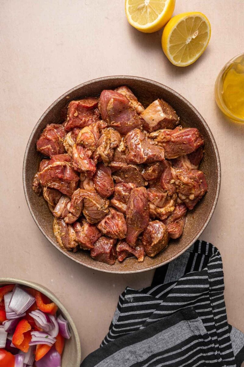 A bowl of marinated raw lamb sits on a counter beside a striped cloth, cut lemon halves, a small bowl of chopped onions and peppers, and a glass of oil nearby.