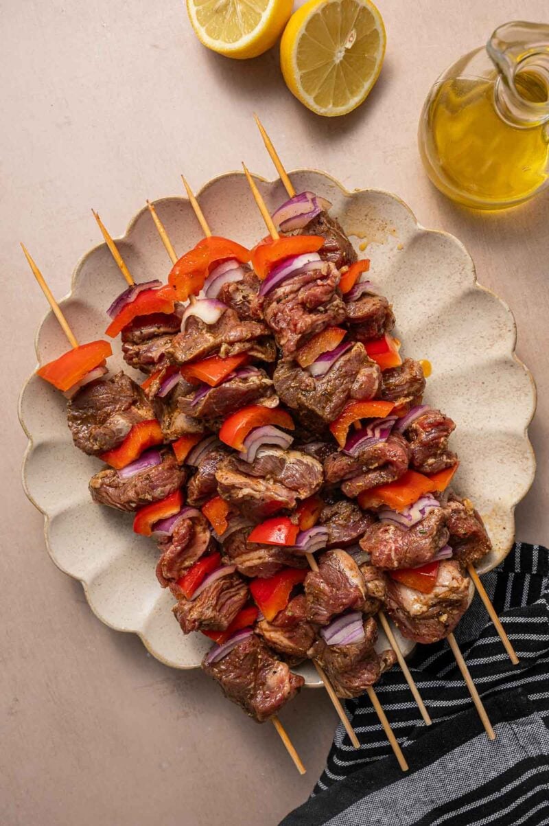 Wooden skewers with marinated lamb, red bell peppers, and red onions arranged on a scalloped plate, with lemon halves and a glass jug of olive oil nearby.