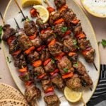 A platter of grilled lamb and vegetable skewers garnished with parsley and lemon wedges, accompanied by flatbread and a bowl of tzatziki sauce.