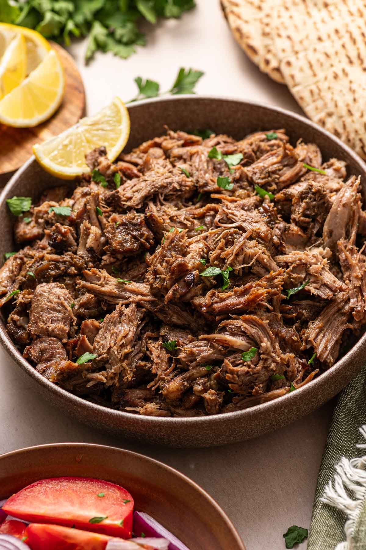A bowl of shredded, seasoned lamb garnished with chopped herbs, with lemon wedges, flatbread, and fresh vegetables nearby.