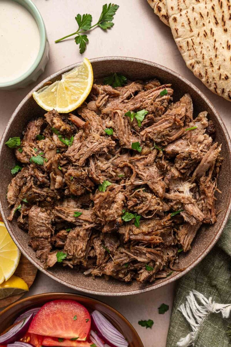 A bowl of shredded, seasoned lamb garnished with parsley, served with a lemon wedge. Pita bread, sauce, and sliced vegetables are nearby.