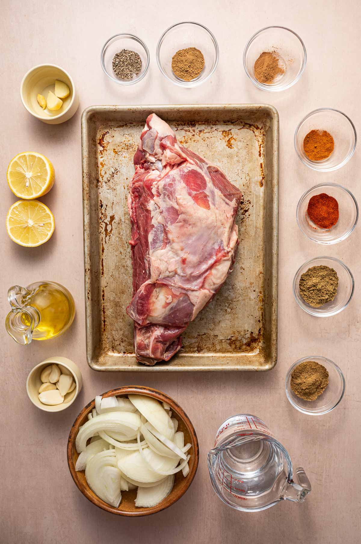 Raw lamb shoulder on a baking tray surrounded by small bowls of spices, sliced onions, garlic cloves, lemon halves, olive oil, and a measuring cup of water on a beige surface.