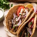 Two lamb shawarma wraps filled with shredded meat, sliced red onions, tomato, fresh parsley, and a drizzle of white sauce, served on a plate with lemon wedges.