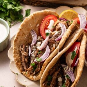 Two lamb shawarma wraps filled with shredded meat, sliced red onions, tomato, fresh parsley, and a drizzle of white sauce, served on a plate with lemon wedges.