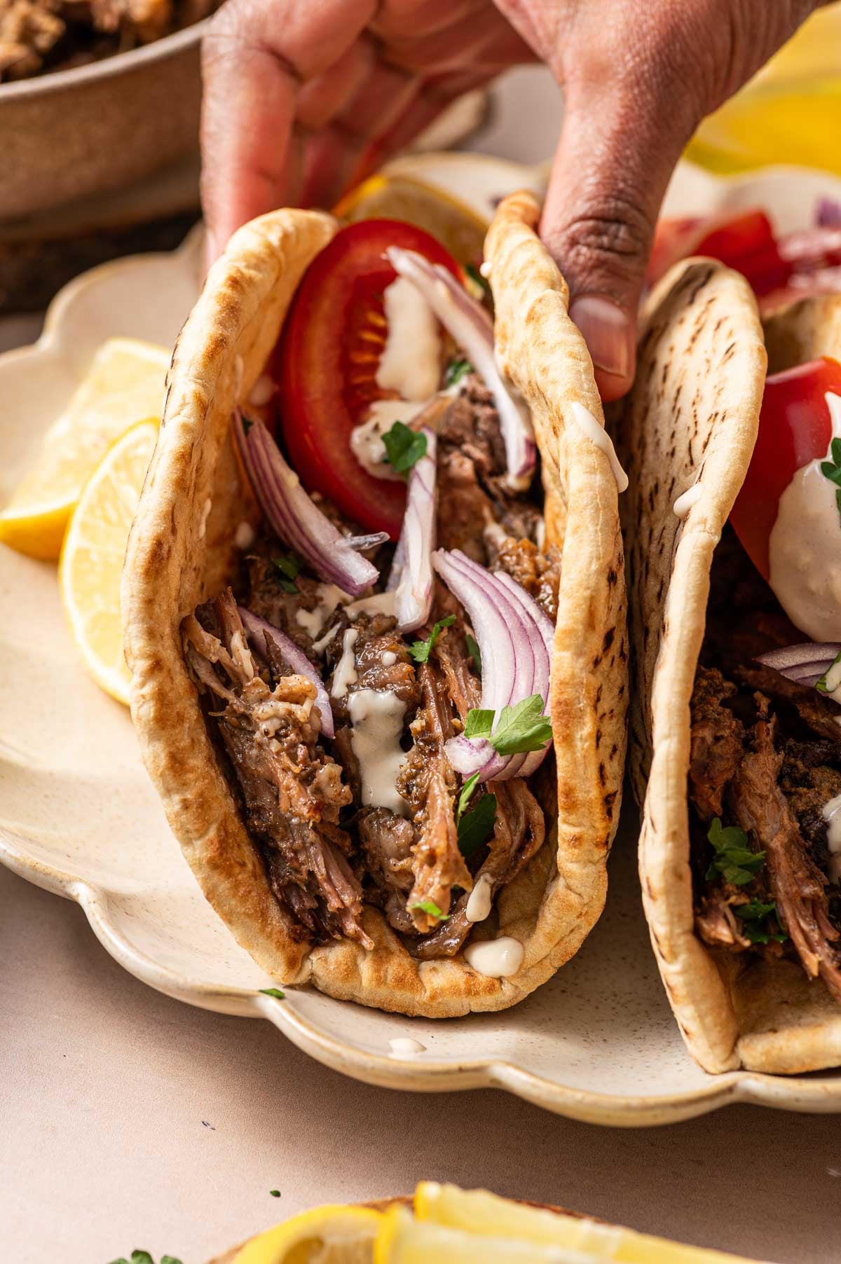 A hand holds a pita stuffed with shredded meat, sliced red onions, tomato, herbs, and a drizzle of white sauce, served on a plate with lemon wedges.