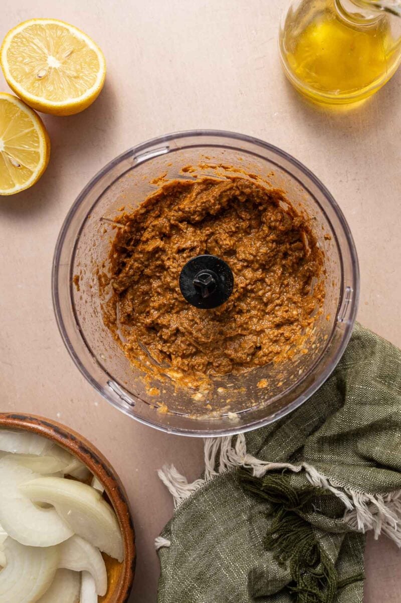 A food processor containing a brown paste sits on a counter, surrounded by a halved lemon, a glass bottle of oil, sliced onions in a bowl, and a green cloth.