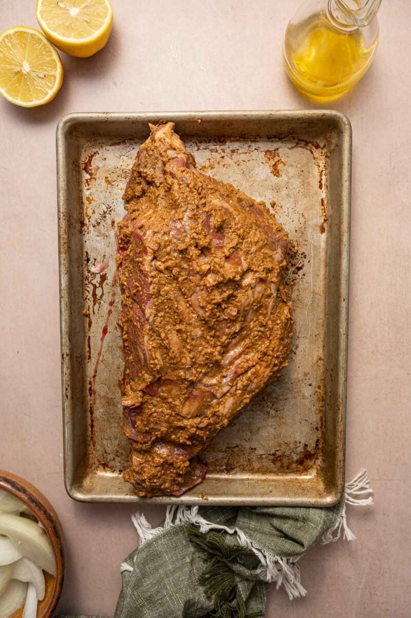 A marinated cut of lamb on a metal baking tray, with a halved lemon, a glass bottle of oil, and a bowl of sliced onions nearby.