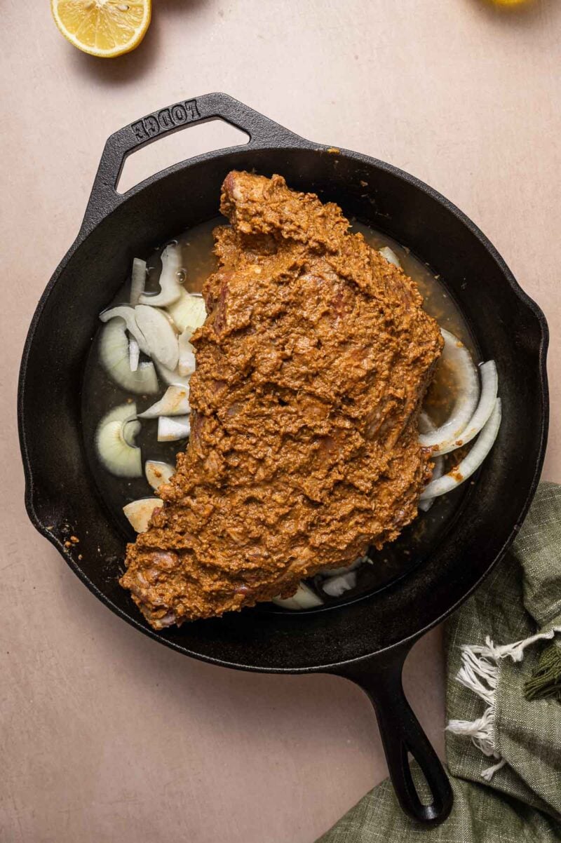 A cast iron skillet with marinated lamb shoulder; coated in a reddish-brown spice paste, resting on sliced onions, ready to be cooked.
