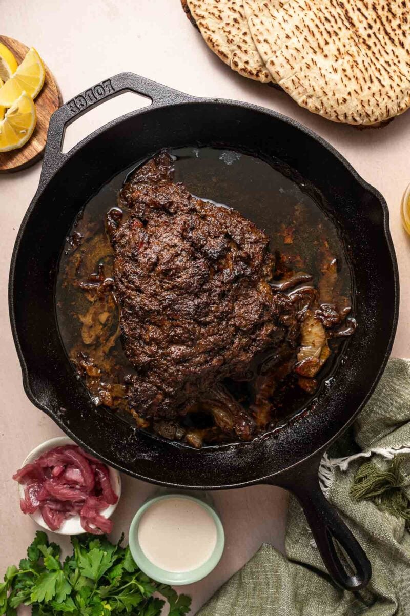 A cast iron skillet with a cooked, seasoned lamb shoulder, surrounded by flatbread, lemon wedges, pickled onions, cilantro, and a bowl of sauce.