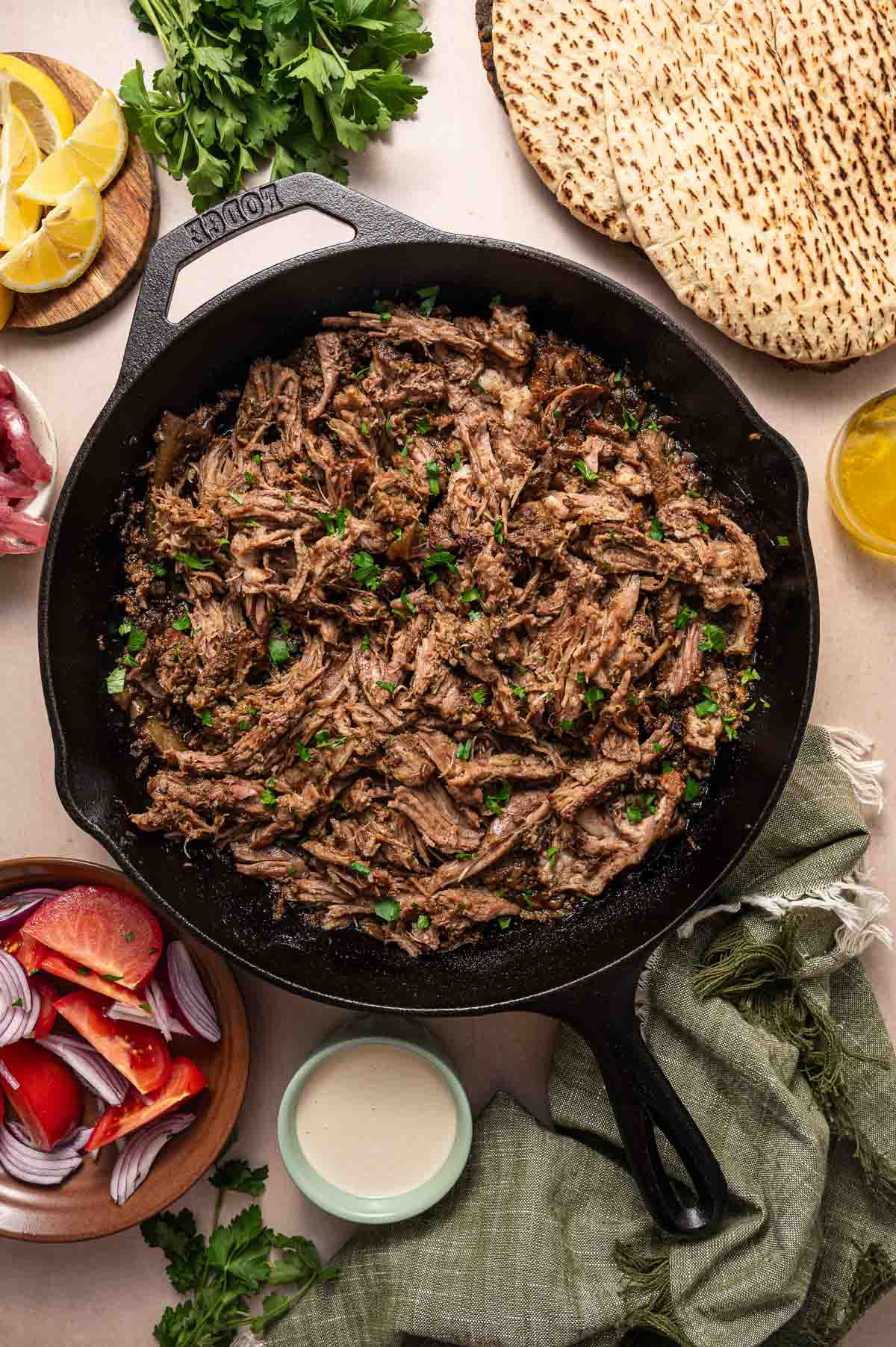 Shredded cooked beef in a cast iron skillet, surrounded by flatbread, lemon wedges, fresh herbs, cut tomatoes, red onions, and a small bowl of sauce.