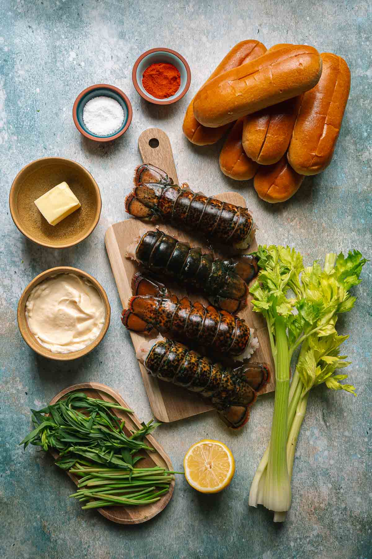 Lobster tails on a cutting board, with hot dog buns, celery, lemon, butter, mayonnaise, tarragon, and spices arranged around them on a textured surface.