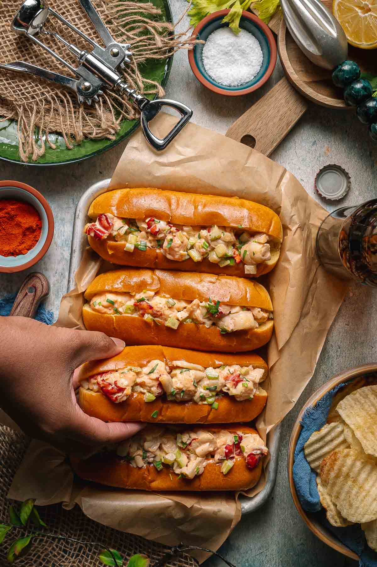Four main lobster rolls on a tray lined with parchment paper, with a hand reaching for one. Chips, seasoning, salt, and lemon are arranged around the tray.