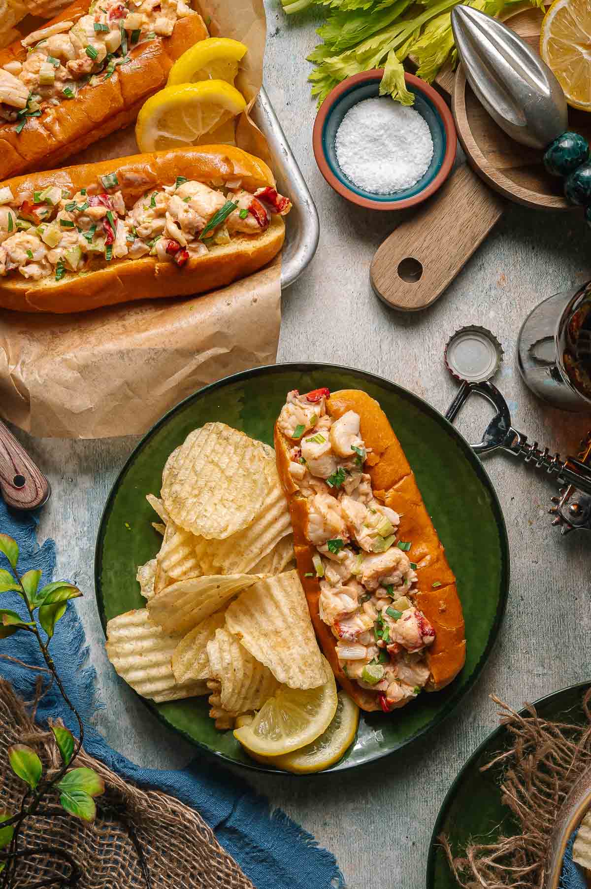 A plate with a Maine lobster roll in a bun, topped with herbs, served with potato chips and lemon slices; more lobster rolls and lemon wedges are on a tray nearby.