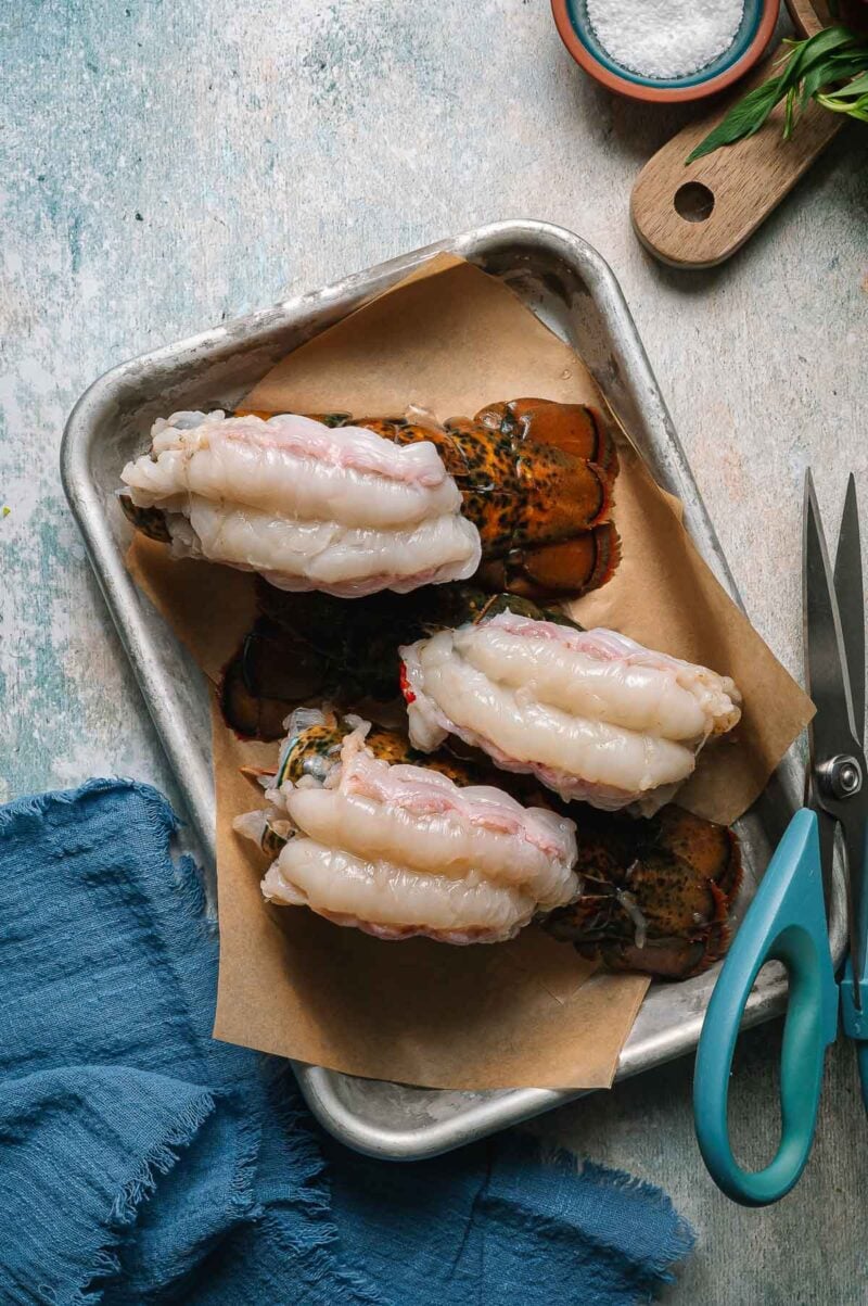 Three raw lobster tails with shells split and meat exposed, arranged on brown parchment paper on a metal tray, with kitchen shears and salt nearby.
