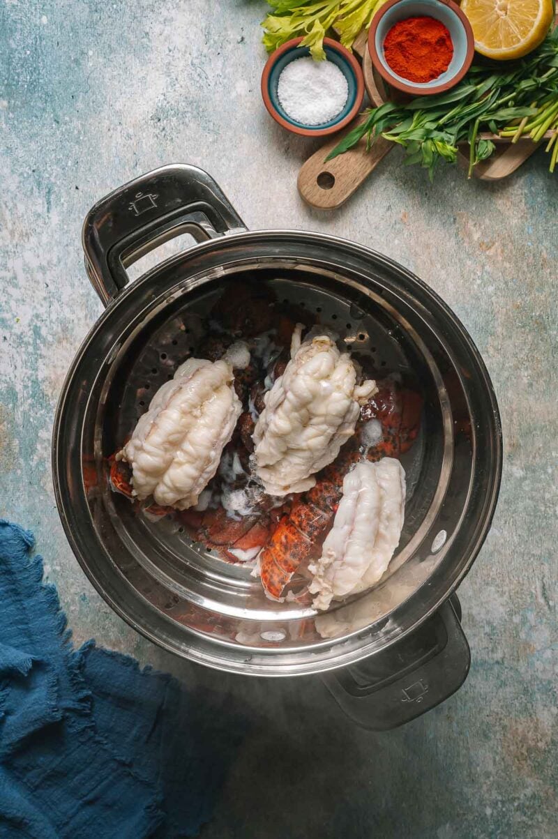 Three lobster tails are being steamed in a pot, surrounded by ingredients such as salt, paprika, fresh herbs, and half a lemon on a nearby cutting board.