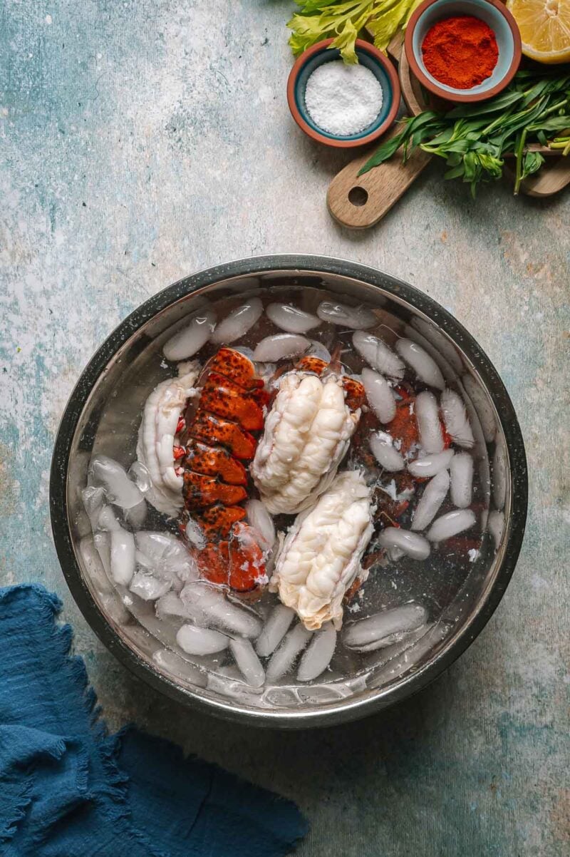 Three lobster tails in a metal bowl of ice water, with seasoning, lemon, and herbs on a cutting board nearby.