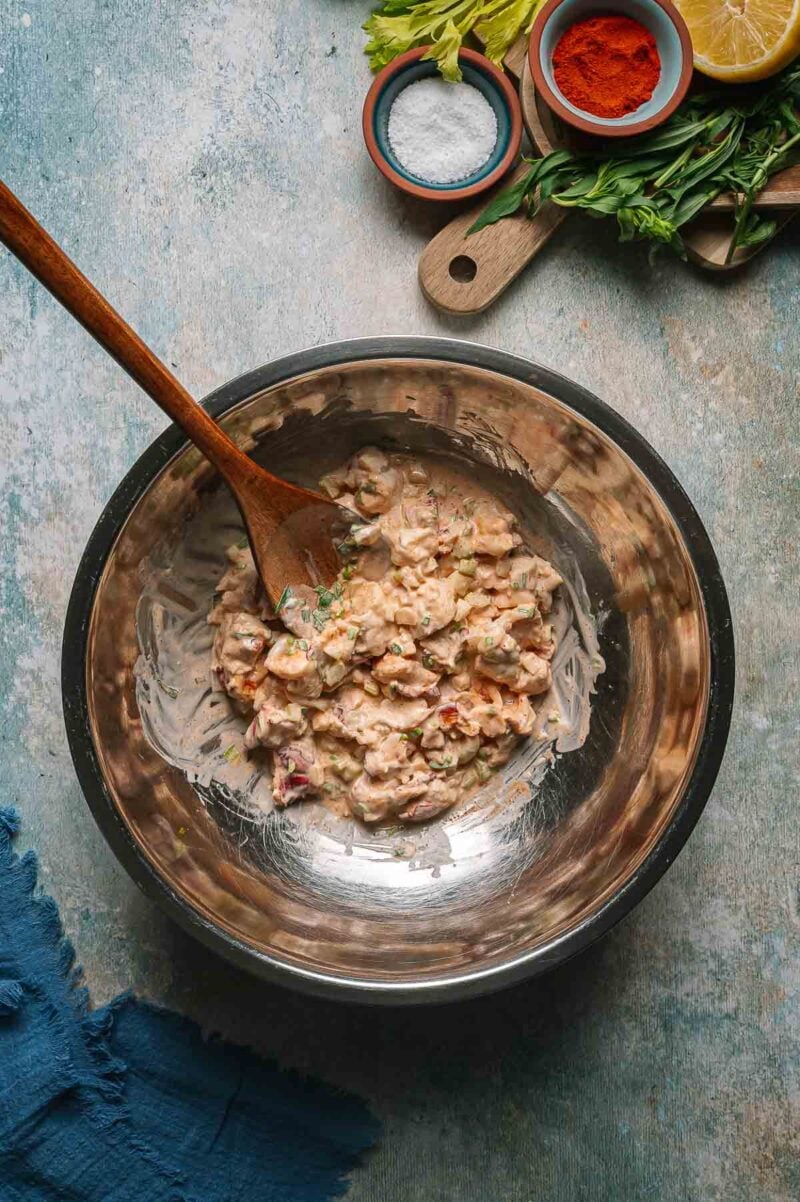 A metal bowl with a creamy lobster salad mixture and wooden spoon, surrounded by herbs, lemon, salt, and paprika on a textured surface.